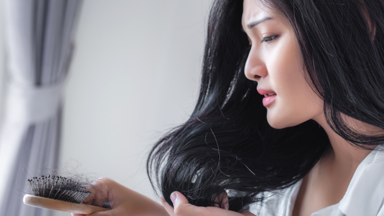 Hispanic woman holding wooden hair brush with a worried face as she stares at clumps of hair in the brush